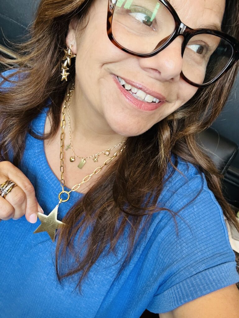 Lori Kappler wearing layered gold necklaces, earrings, and rings while showcasing personalized jewelry from Blooming with Lori.