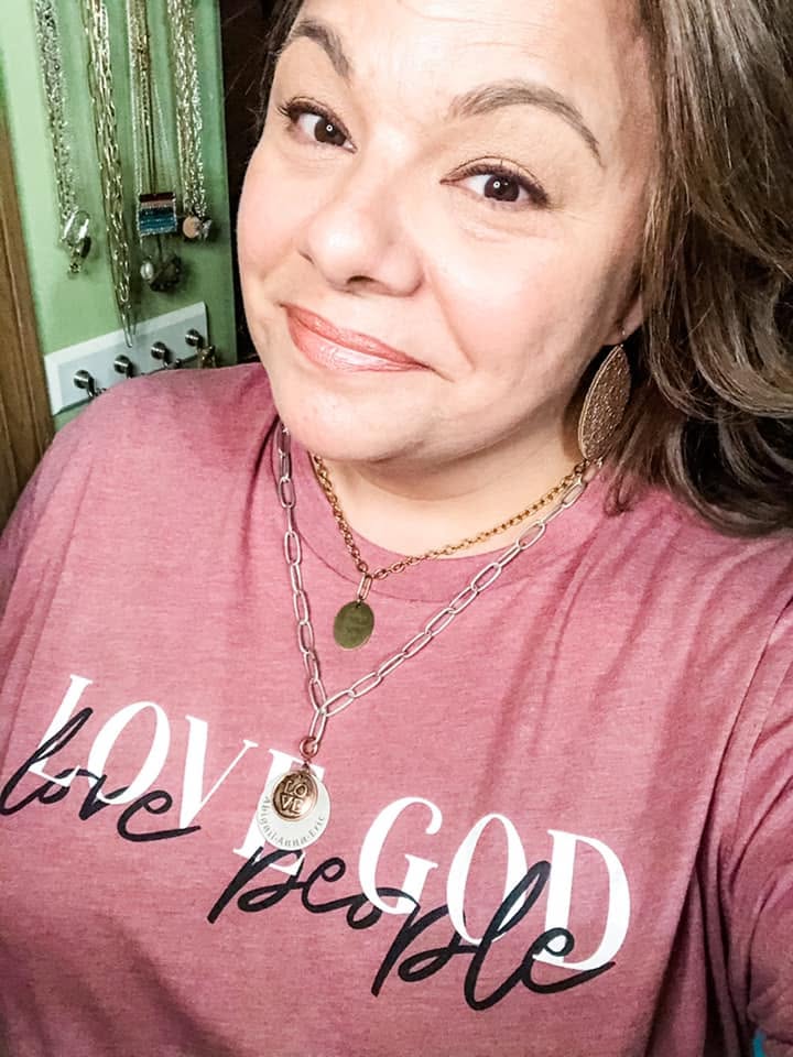 Lori smiling in a Love God Love People shirt, wearing her personalized necklaces engraved with her kids' names